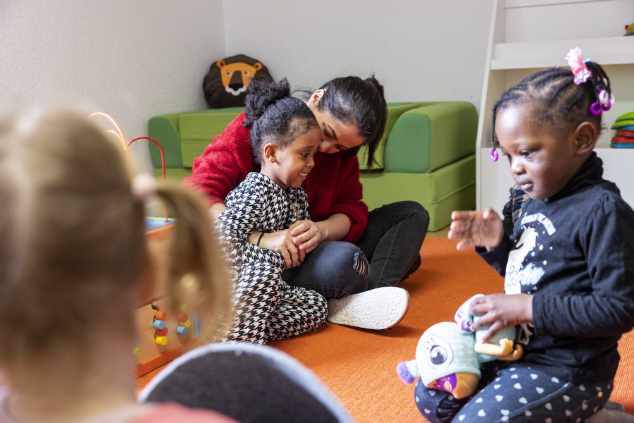 Zwei Kinder und eine erwachsene Person sitzen auf dem Boden, die erwachsene Person umarmt das eine Kind, das andere Kind spielt mit einem Spielzeug