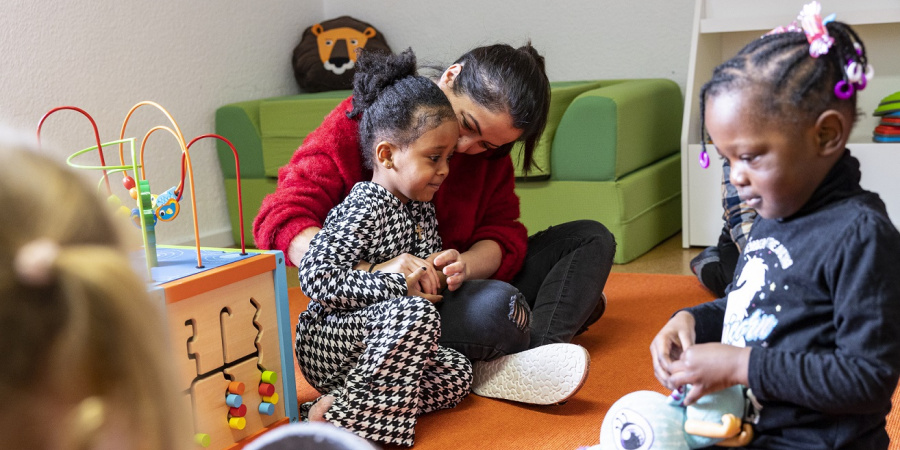 Zwei Mädchen sitzen auf dem Boden. Das eine Mädchen wird von einer Frau umarmt, das andere spielt mit einem Kuscheltier.