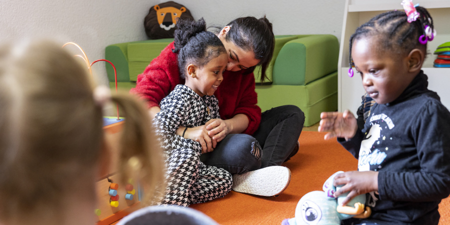 Zwei Kinder und eine erwachsene Person sitzen auf dem Boden, die erwachsene Person umarmt das eine Kind, das andere Kind spielt mit einem Spielzeug