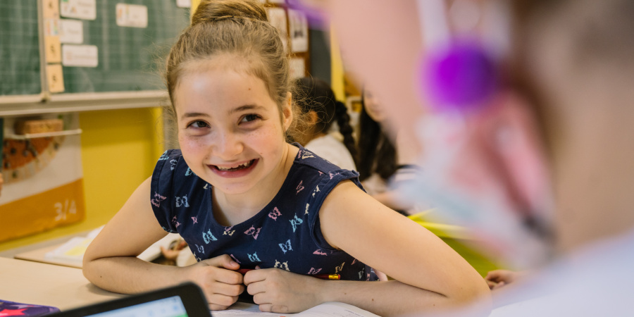 Ein Mädchen sitzt am Schultisch und schaut lachend ihre MItschülerin an.